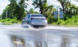 A car driving through a flooded street with water splashing on both sides, surrounded by green trees and road signs, highlighting the importance of flood elevation surveys for assessing flood risks in residential areas.