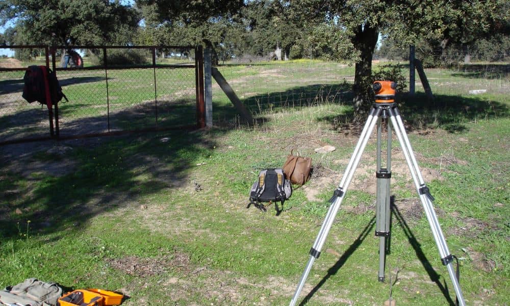 Land Surveying Equipment on a Residential Property Site - ALTA SURVEY Florida Tripod-mounted land surveying instrument set up by a land surveyor near a fence on a grassy residential property in Jacksonville, Florida, used for marking property lines before home improvements.